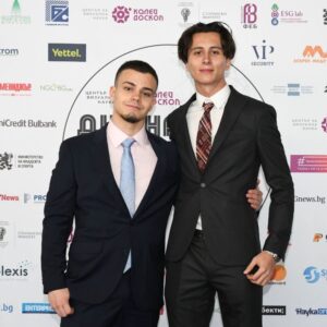 Двама млади мъже, облечени официално, позират на фона на стена с логотипи на партньори и спонсори по време на събитие. Two young men in formal attire posing in front of a backdrop with partner and sponsor logos during an event.