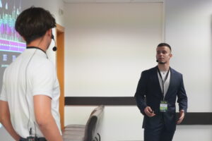 Двама млади мъже с микрофони провеждат презентация в професионална обстановка, като един от тях е облечен в костюм. Two young men with microphones delivering a presentation in a professional setting, one dressed in a suit.