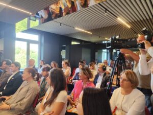 Зала с хора, седящи и слушащи презентация, докато журналисти с камери документират събитието. A room filled with people sitting and listening to a presentation while journalists with cameras document the event.